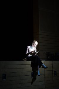 Eine Person mit blonden Haaren sitzt auf einer erhöhten Fläche und liest ein Buch. Sie ist von einem Lichtstrahl beleuchtet, während der Hintergrund dunkel ist. Die Person trägt einen eleganten, hellen Oberteil und schwarze Hosen.