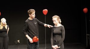 Drei Personen in einem Theaterraum mit dunklem Hintergrund. Zwei Personen tragen schwarze Kleidung, eine von ihnen hält einen roten Ballon. Eine der Personen steht direkt vor der anderen, die den Kopf gesenkt hat. Im Hintergrund ist eine weitere Person mit einer weißen Maske zu sehen.