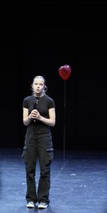 Eine junge Frau steht auf einer Bühne mit einem Mikrofon in der Hand. Sie trägt ein schwarzes T-Shirt und schwarze Hosen. Im Hintergrund schwebt ein roter Luftballon. Der Bühnenboden ist dunkel und die Beleuchtung konzentriert sich auf die Frau.