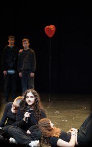 Eine Gruppe von Jugendlichen auf einer Theaterbühne. Eine junge Frau sitzt im Vordergrund und hält ein Mikrofon, während sie spricht. Um sie herum liegen andere Jugendliche in schwarzen Outfits. Im Hintergrund stehen zwei weitere Jugendliche, die ebenfalls in dunkler Kleidung gekleidet sind. Ein rotes Herz-Ballon schwebt hinter ihnen. Die Szene vermittelt eine emotionale und dramatische Atmosphäre.