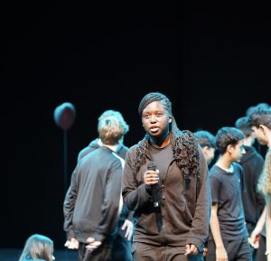 Ein junges Mädchen mit langen, lockigen Haaren steht auf einer Bühne und hält ein Mikrofon. Sie trägt ein schwarzes Outfit und spricht oder singt. Im Hintergrund sind mehrere Personen in schwarzen Kleidungsstücken, die sich auf der Bühne versammeln. Es ist eine Aufführung oder ein Gesangsauftritt zu sehen.