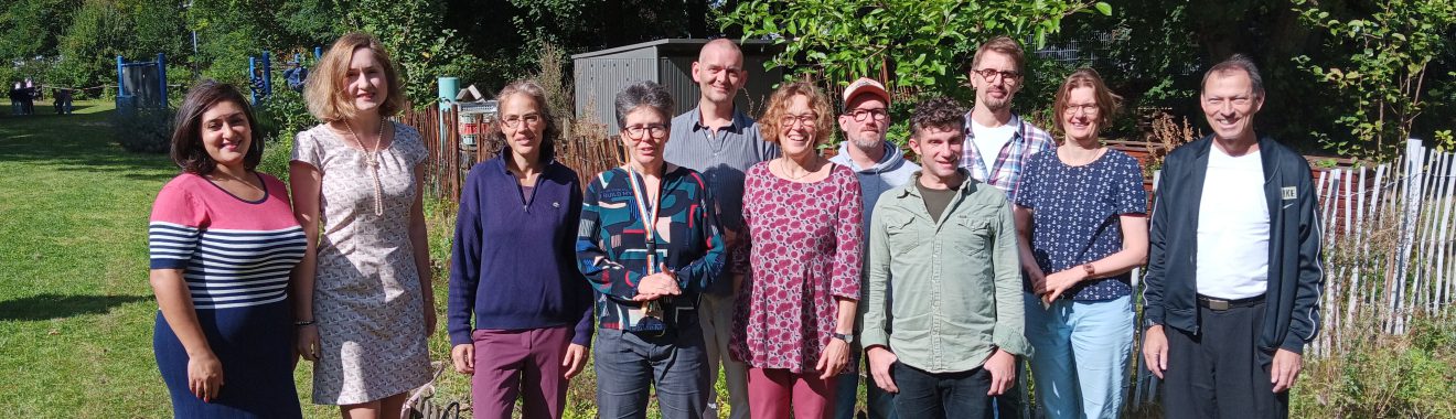 Eine Gruppe von 13 Personen steht auf einer Wiese, umgeben von Bäumen. Die Menschen lächeln und schauen in die Kamera. Sie tragen verschiedene, sommerliche Kleidung. Im Hintergrund sind einige grüne Pflanzen und ein Zaun sichtbar.