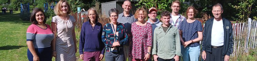 Eine Gruppe von 13 Personen steht auf einer Wiese, umgeben von Bäumen. Die Menschen lächeln und schauen in die Kamera. Sie tragen verschiedene, sommerliche Kleidung. Im Hintergrund sind einige grüne Pflanzen und ein Zaun sichtbar.