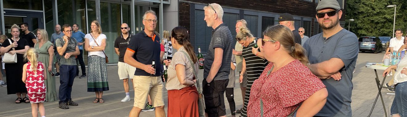 Eine Gruppe von Menschen steht vor einem Gebäude, einige sind in einer Schlange. Es sind verschiedene Altersgruppen und Kleidungsstile vertreten. Im Vordergrund stehen mehrere Personen, während weitere im Hintergrund sichtbar sind. Es ist Tageslicht und die Atmosphäre wirkt entspannt.