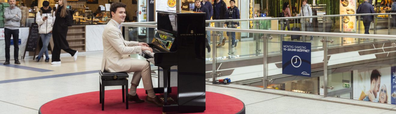 Ein Pianist sitzt an einem Klavier auf einem roten Teppich in einem Einkaufszentrum. Um ihn herum stehen Menschen, die ihm zuschauen. Im Hintergrund sind Geschäfte und eine offene Galerie sichtbar.