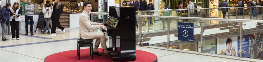 Ein Pianist sitzt an einem Klavier auf einem roten Teppich in einem Einkaufszentrum. Um ihn herum stehen Menschen, die ihm zuschauen. Im Hintergrund sind Geschäfte und eine offene Galerie sichtbar.