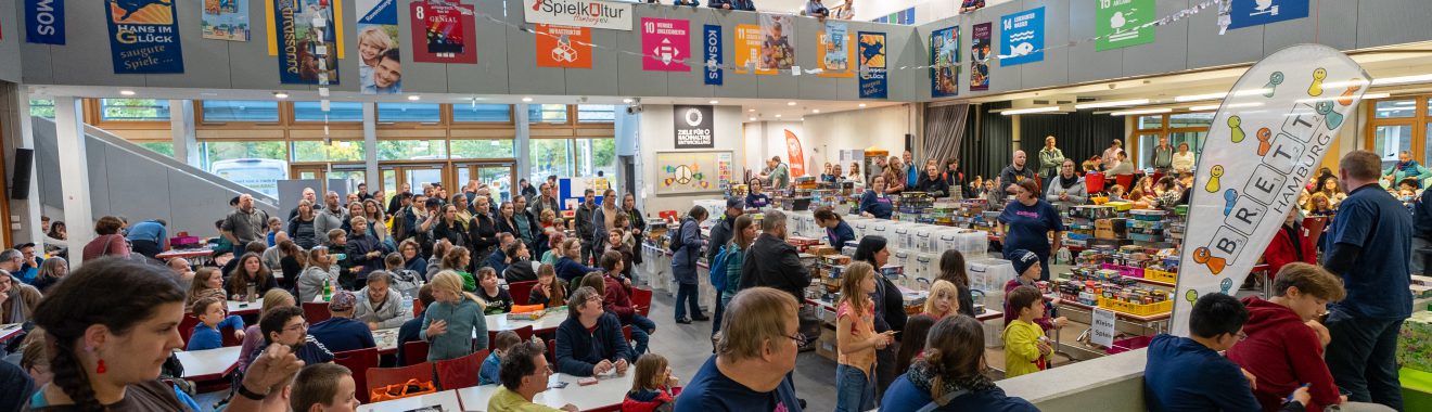 Eine große Gruppe von Menschen sitzt in einem modernen Veranstaltungsraum. Im Hintergrund sind auf verschiedenen Ebenen Banner und Plakate sichtbar. Die Anordnung der Sitzplätze ist offen, und die Atmosphäre wirkt lebhaft und einladend. Viele Personen sind in Gespräche vertieft oder hören aufmerksam zu.