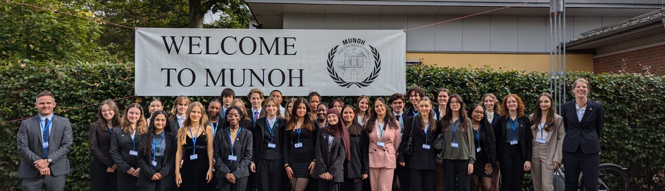 Eine Gruppe von etwa 40 Personen steht vor einem großen Banner mit der Aufschrift "WELCOME TO MUNOAH". Die Personen tragen formelle Kleidung und lächeln in die Kamera. Im Hintergrund sind Bäume und ein Gebäude sichtbar, der Himmel ist teilweise bewölkt.