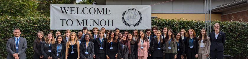 Eine Gruppe von etwa 40 Personen steht vor einem großen Banner mit der Aufschrift "WELCOME TO MUNOAH". Die Personen tragen formelle Kleidung und lächeln in die Kamera. Im Hintergrund sind Bäume und ein Gebäude sichtbar, der Himmel ist teilweise bewölkt.