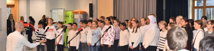 Ein Kinder- und Jugendchor steht auf einer Bühne und singt. Die Mitglieder tragen einheitliche Shirts in Schwarz und Weiß. Vor dem Chor steht ein Dirigent, der sie anleitet. Im Hintergrund sind verschiedene farbige Banner angebracht, und im Vordergrund sind Tische mit Getränken und persönlichen Gegenständen zu sehen. Die Szene vermittelt eine lebhafte und musikalische Atmosphäre.