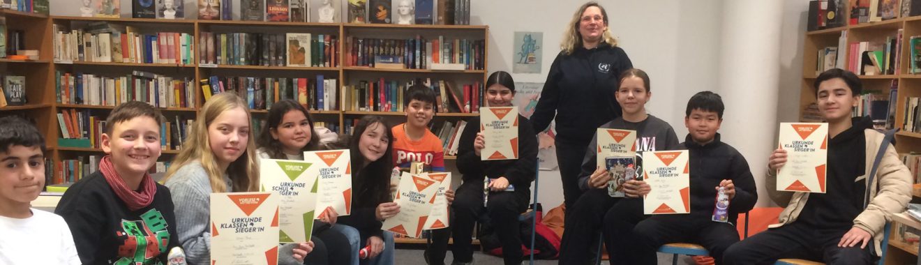 Eine Gruppe von zehn Jugendlichen sitzt und steht in einer Bibliothek. Sie halten Urkunden oder Zertifikate in den Händen. Im Hintergrund sind Regale mit vielen Büchern zu sehen. Die Atmosphäre wirkt freundlich und feierlich.