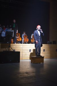 Ein Mann steht auf einer Kiste und singt in ein Mikrofon, während er von einem Orchester begleitet wird. Im Hintergrund sind Musiker mit Celli zu sehen. Der Mann trägt einen schwarzen Anzug und eine rote Mütze. Die Bühne ist schwach beleuchtet, was eine dramatische Atmosphäre schafft.