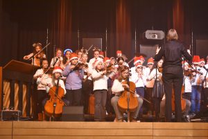 Ein Kinderorchester steht auf der Bühne und spielt. Die Musiker tragen Weihnachtsmützen und spielen verschiedene Instrumente wie Violine und Cello. Ein Dirigent leitet das Orchester von vorne. Die Bühne ist festlich beleuchtet.