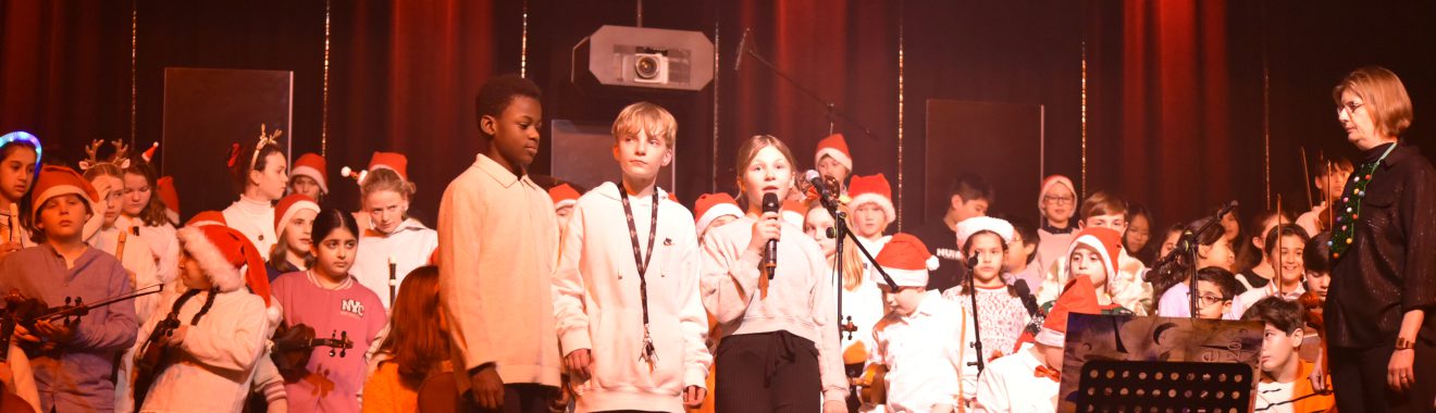 Eine Gruppe von Schülern steht auf der Bühne und singt. Viele tragen Weihnachtsmützen und weiße Hemden. Einige halten Instrumente. Die Bühne ist mit bunten Lichtern beleuchtet, und im Hintergrund sind weitere Schüler zu sehen. Eine Lehrkraft steht rechts und schaut auf die Gruppe.