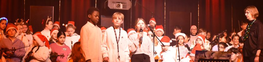 Eine Gruppe von Schülern steht auf der Bühne und singt. Viele tragen Weihnachtsmützen und weiße Hemden. Einige halten Instrumente. Die Bühne ist mit bunten Lichtern beleuchtet, und im Hintergrund sind weitere Schüler zu sehen. Eine Lehrkraft steht rechts und schaut auf die Gruppe.