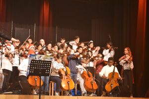 Eine Gruppe von Kindern und Jugendlichen spielt auf der Bühne verschiedene Musikinstrumente, darunter Geigen und Celli. Im Hintergrund sind einige Musiker zu sehen, die konzentriert musizieren. Die Bühne ist schwach beleuchtet, mit rotem Licht, und ein Notenständer steht im Vordergrund.