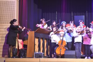 Eine Gruppe von jungen Musikern spielt auf der Bühne, während eine Lehrerin mit einem Geigenbogen dirigiert. Die Kinder tragen Weihnachtsmützen und spielen verschiedene Streichinstrumente, darunter Geigen und ein Cello. Im Hintergrund sind bunte Lichter und eine festliche Dekoration zu sehen.