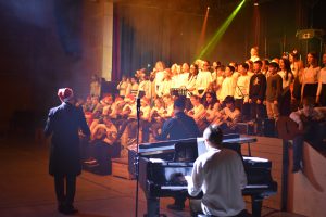 Eine Gruppe von Kindern und Jugendlichen steht auf einer Bühne und singt. Im Vordergrund sitzen zwei Musiker an einem Klavier. Der Raum ist schwach beleuchtet, mit bunten Lichtstrahlen, die von der Bühne ausgehen. Die Sänger tragen überwiegend helle Kleidung, während einer der Musiker einen schwarzen Anzug und einen Hut trägt.