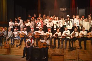 Eine Gruppe von Kindern und Jugendlichen steht und sitzt auf einer Bühne, die musizieren und singen. Viele tragen Weihnachtsmützen und T-Shirts. Einige halten Gitarren, während andere im Hintergrund stehen und mitspielen. Die Bühne ist festlich dekoriert, und es gibt ein leichtes Nebellicht.