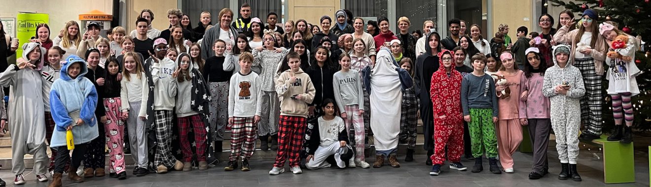 Gruppenfoto von mehreren Personen, einschließlich Kinder und Erwachsene, die in festlichen Schlafanzügen gekleidet sind. Sie stehen in einer Halle, umgeben von weihnachtlicher Dekoration, darunter ein Weihnachtsbaum. Alle Personen lächeln und posieren gemeinsam für das Bild.