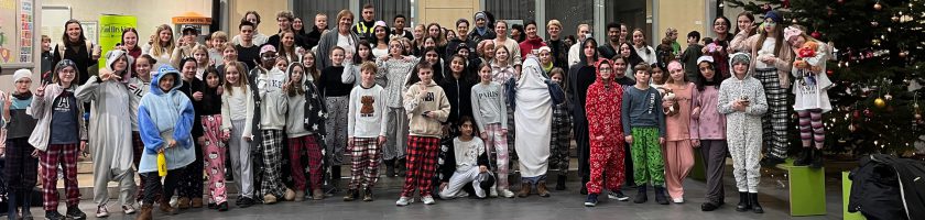 Gruppenfoto von mehreren Personen, einschließlich Kinder und Erwachsene, die in festlichen Schlafanzügen gekleidet sind. Sie stehen in einer Halle, umgeben von weihnachtlicher Dekoration, darunter ein Weihnachtsbaum. Alle Personen lächeln und posieren gemeinsam für das Bild.