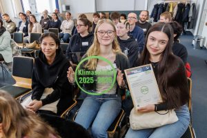 Drei junge Frauen sitzen in einem Publikum, halten ein Schild mit der Aufschrift „Klimaschutz 2025“ und eine Urkunde. Im Hintergrund sind weitere Personen in einem Raum zu sehen.