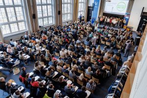 Eine große Veranstaltung in einem modernen Saal mit vielen Sitzplätzen. Im Vordergrund stehen mehrere Personen auf einer Bühne, während im Hintergrund zahlreiche Zuhörer in Reihen sitzen und auf die Bühne schauen. Große Fenster lassen Tageslicht in den Raum.