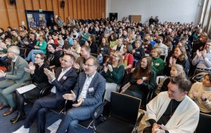 Eine große Gruppe von Menschen sitzt in einem Raum und applaudiert. Die Zuschauer sind vielfältig gekleidet und zeigen positive Reaktionen. Im Hintergrund sind weitere Personen und eine Bühne zu sehen. Die Atmosphäre ist lebhaft und engagiert.