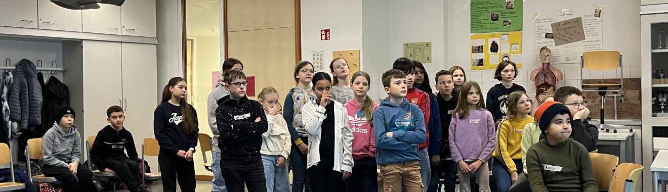 Eine Gruppe von Kindern und Jugendlichen steht in einem Klassenzimmer. Im Vordergrund sind einige Stühle und Tische sichtbar. Die Schüler sind in verschiedenen Positionen angeordnet und tragen unterschiedliche Kleidung. Die Wände des Raumes sind teilweise mit Postern und Materialien dekoriert.