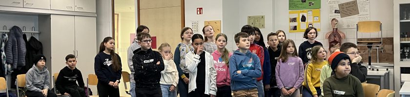 Eine Gruppe von Kindern und Jugendlichen steht in einem Klassenzimmer. Im Vordergrund sind einige Stühle und Tische sichtbar. Die Schüler sind in verschiedenen Positionen angeordnet und tragen unterschiedliche Kleidung. Die Wände des Raumes sind teilweise mit Postern und Materialien dekoriert.