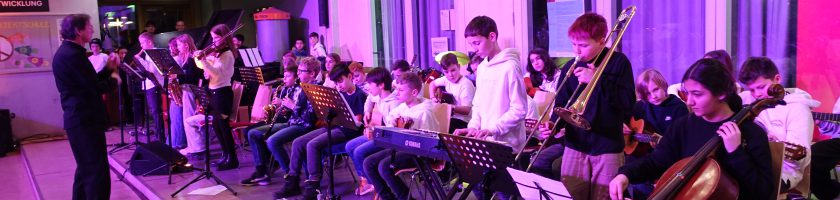 Eine Gruppe von Musikern spielt in einem Innenraum. Die Musiker sind in zwei Reihen aufgestellt, einige spielen auf Streichinstrumenten, während andere Blasinstrumente und Percussion verwenden. Im Hintergrund ist eine Lehrperson zu sehen, die die Gruppe anleitet. Der Raum ist mit bunten Lichtern beleuchtet und es hängen Plakate an den Wänden.