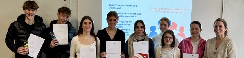 Eine Gruppe von neun Personen steht zusammen und hält jeweils ein Dokument in der Hand. Sie lächeln und posieren vor einer Wand, die teilweise mit grafischen Elementen und Texten gestaltet ist. Die Personen sind unterschiedlichen Alters und Geschlechts. Einige tragen lässige Kleidung, während andere etwas formeller gekleidet sind.