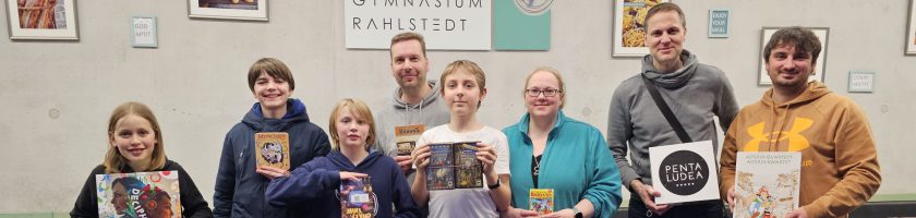 Gruppe von sieben Personen steht lächelnd vor einer Wand mit Kunstwerken. Sie halten verschiedene Bücher in den Händen. Im Hintergrund sind einige Bilder zu sehen, sowie ein Schild mit dem Text "SPANNSTOWN FAMILIERT".