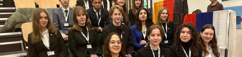 Gruppensfoto von mehreren jungen Erwachsenen, die in formeller Kleidung gekleidet sind. Sie stehen und sitzen auf einer Treppe und lächeln in die Kamera. Im Hintergrund sind farbige Flaggen und Kunstwerke zu sehen.